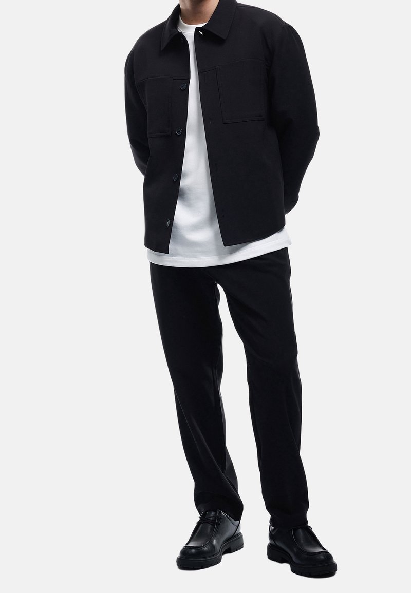 Un homme porte une veste noire boutonnée, un t-shirt blanc, un pantalon noir et des chaussures noires, debout avec les mains derrière le dos sur un fond blanc.