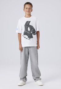 Balts kokvilnas T-krekls ar lielu melnu grafiku stilizētam trušim, apvienots ar brīviem pelēkiem džinsa biksēm un baltām sporta apaviem.