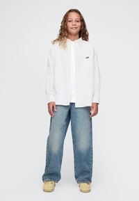 Vaikas su garbanotais plaukais, vilkintis per didelę baltą marškinių su „GAP“ logotipu, laisvus mėlynus džinsus ir smėlio spalvos batus, stovintis prie vienspalvio fono.