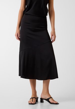 Femme portant une jupe midi noire avec une couture asymétrique, associée à des sandales noires à talons blocs et bouts ouverts, debout sur un sol blanc.