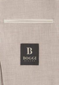 Tissu à petits carreaux beige et blanc avec une étiquette noire cousue comportant un grand "B" et le texte "Boggi Milano" en dessous.
