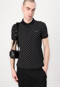 Svart polo skjorte med et geometrisk mønster i grått, korte ermer og svart krage. Modellen bærer også en liten svart veske.