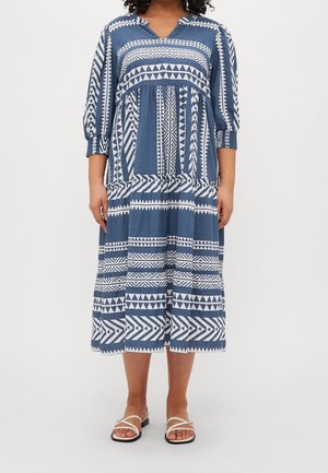 Woman wearing a blue and white geometric patterned midi dress with 3/4 sleeves and white flat sandals.