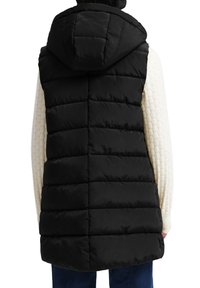 Veste noire matelassée sans manches avec capuche portée sur un haut blanc texturé à manches longues et un pantalon bleu foncé, vue de dos.