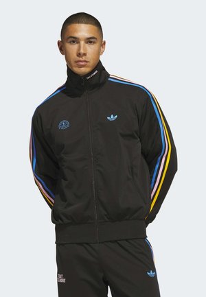 Mand iført en sort Adidas træningsdragt med flerfarvede striber på ærmer og bukser, stående med hænderne bag ryggen.