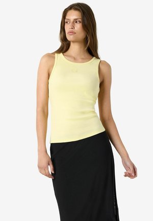 Femme aux longs cheveux bruns portant un haut sans manches jaune pâle et une jupe noire, regardant vers sa gauche sur un fond clair uni.