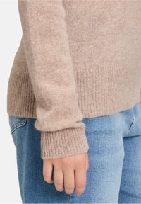 Jasno beżowy sweter wykonany na drutach z prążkowanymi mankietami, uszyty z miękkiego materiału, zestawiony z niebieskimi dżinsami. Skupienie na fakturze i kontraście kolorów.