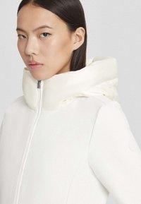 Giovane donna con capelli castani lisci che indossa un giacca bianca con colletto alto e cappuccio imbottito, rivolta leggermente a sinistra su uno sfondo neutro.