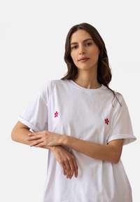 Biała bawełniana koszulka z krótkim rękawem, z dwoma małymi różowymi haftami kwiatowymi na piersi. Miękka tekstura i luźny krój.