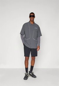 Hall halli lühikeste varrukatega nööbitav särk valgete aksentidega, kombineeritud mustade lühikeste pükste ja tumedate tossudega, kantud päikeseprillide ja ülekattevarrukatega.