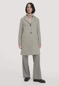 Grauer Wollmantel mit Reverskragen, vorderer Knopfleiste und Seitentaschen. Darunter ein rosa Strickpullover und graue Hosen mit weitem Bein.