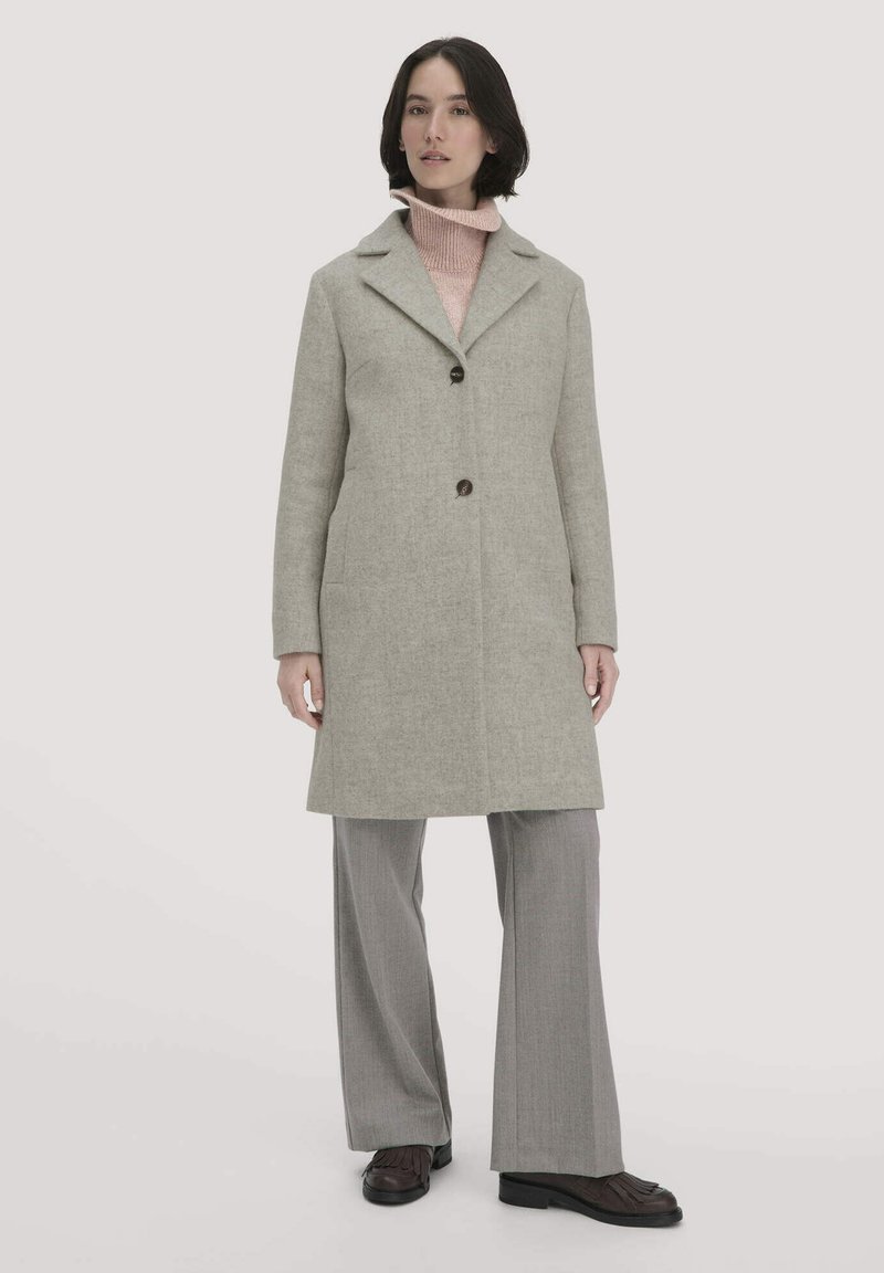 Grauer Wollmantel mit Reverskragen, vorderer Knopfleiste und Seitentaschen. Darunter ein rosa Strickpullover und graue Hosen mit weitem Bein.