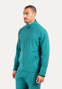 Teal Zip-Jacke aus weichem Stoff mit Stehkragen und seitlichen Reißverschlusstaschen; glatte Textur mit figurbetontem Design.