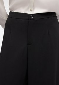 Pantaloni neri su misura con una superficie liscia, caratterizzati da un taglio dritto, pieghe frontali e una chiusura con un singolo bottone nero in vita.