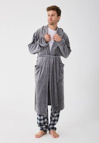 Man standing barefoot wearing a gray plush robe over a white shirt and black-and-white checkered pajama pants, holding the robe collar.