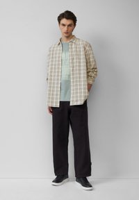 Button-up shirt in crème met een beige ruitpatroon, gedragen over een mintgroen graphic t-shirt. Zwarte wijd-uitlopende broek en zwarte sneakers.