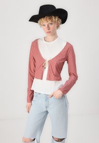 Piros és fehér kockás cropped kardigán hosszú ujjal, elöl megkötős záródással, fehér ing fölé rétegezve, világoskék elhasználódott sorttal párosítva.