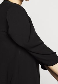 Svart topp med trekvartsärmar. Slät textur, minimalistisk design, bred halsringning och subtil sömdetaljering längs ärmarna.