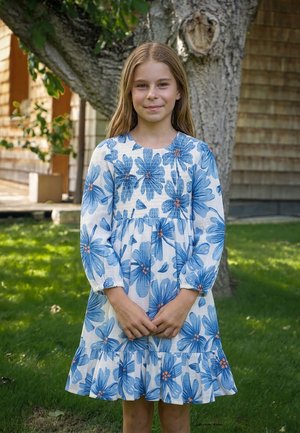 Junges Mädchen mit langen blonden Haaren steht im Freien und trägt ein weißes Kleid mit großem blauem Blumenmuster, die Hände vor dem Körper verschränkt, lächelt sanft.