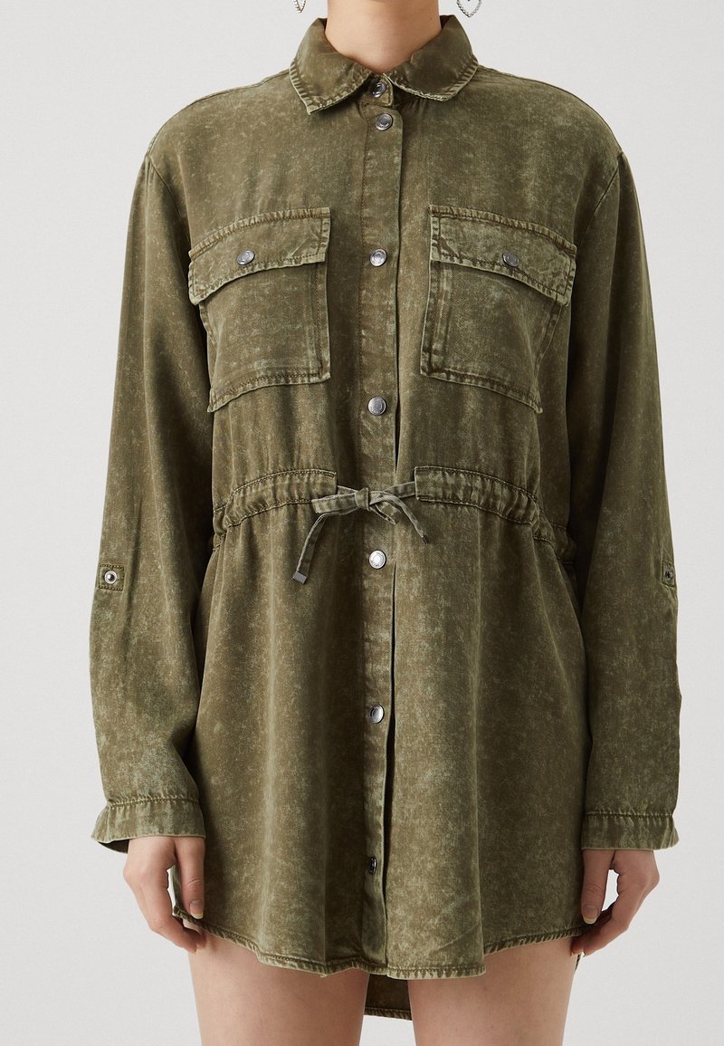 Robe-shirt vert olive en tissu doux, avec un devant à boutons, deux poches poitrine, une taille élastique avec cordon de serrage et des manches longues.