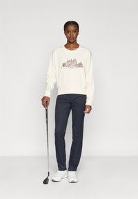 Kreemilonk袖衫，搭配棕色城堡图案，深色裤子和白色运动鞋，手中持着高尔夫球杆。简单设计，休闲合身。