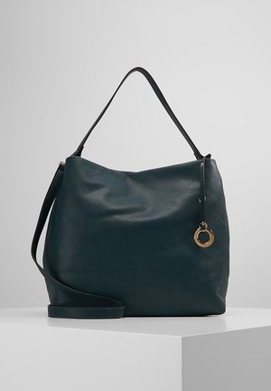 Donkergroene leren schoudertas met een gladde textuur, met een gouden ronde hanger en een verstelbare crossbodyriem op een witte ondergrond.