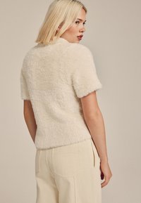 Haut à manches courtes en fausse fourrure crème avec une texture douce et une coupe décontractée, associé à un pantalon beige clair taille haute avec poches latérales.