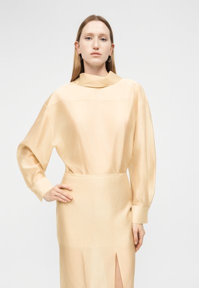 Γυναίκα με ίσια ξανθά μαλλιά, φοράει μακρυμάνικο μπεζ φόρεμα με ψηλό γιακά και πλευρική σχισμή, στέκεται με το χέρι στη μέση.