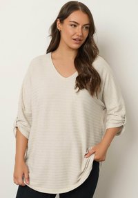 Lichtbeige pullover met een V-hals, lange mouwen en opgerolde manchetten. Heeft een gestructureerd gestreept patroon en zijzakken.