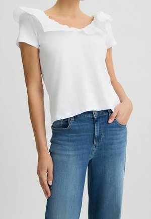 Kobieta w białej bluzce z falbankami i krótkimi rękawami, lekko wpuszczonej w niebieskie dżinsy z wysokim stanem, z jedną ręką w kieszeni.