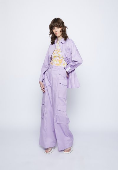 Casaco oversized roxo claro e calças cargo a condizer com grandes bolsos; sobreposto a uma blusa com padrão floral. Textura suave, design casual.