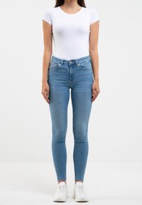 Jeans skinny de color azul claro hechos de denim, con cinco bolsillos, una bragueta con botones y una cintura de altura media, combinados con una camiseta blanca ajustada y zapatillas blancas.