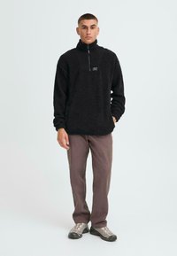 Schwarzer Fleece-Pullover mit hohem Kragen und halbem Reißverschluss. Das Modell trägt locker sitzende graue Hosen und leichte graue Schuhe. Minimale Markenkennzeichnung sichtbar.