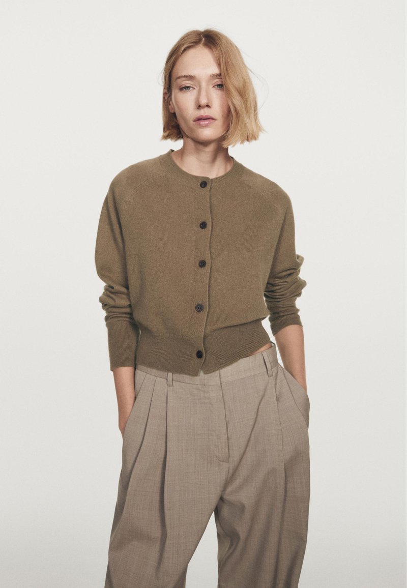 Olive green knitted cardigan with a round neckline, front buttons, and ribbed cuffs, paired with high-waisted beige pleated trousers.
