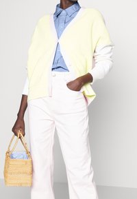 Ljusgul ribbad cardigan med rosa kant, bärs över en blå skjorta; kombinerad med ljusrosa byxor och en vävd brun korgväska.