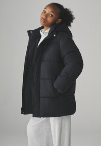 Sort pufferjakke med hætte, præget af en quiltet design, tryklås og sidelommer, båret over et lys gråt beklædningsstykke.