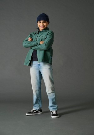 Ung kille står självsäkert med armarna i kors, iklädd en svart beanie, grön jacka, svart tröja, ljusblå jeans och svarta sneakers.