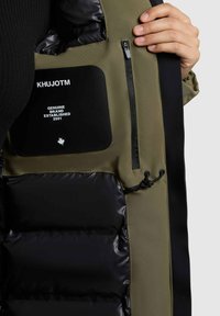 Khaki puchowa kurtka wewnętrzna z czarną podszewką, kieszonką na zamek i naszywką z logo; posiada elastyczny sznur do regulacji i gładką teksturę.