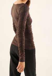 Top marrón de manga larga con textura fruncida en las mangas y los lados, silueta ajustada, utilizado con pantalones negros, diseño minimalista.