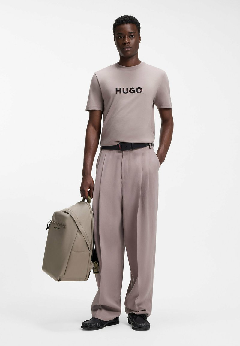 Homme portant un t-shirt beige HUGO et un pantalon plissé, tenant un sac à dos beige assorti, debout devant un fond blanc.