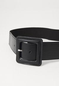 Ceinture en cuir noir avec une boucle carrée, dotée d'une broche en argent et de plusieurs trous pour les ajustements. Texture lisse avec des bords cousus.