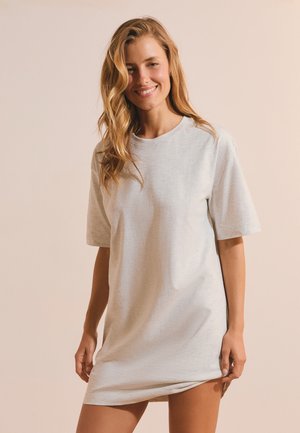 Lachende vrouw met lang blond haar die een lichtgrijze oversized T-shirtjurk draagt, staand tegen een beige achtergrond.