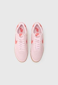 Rosa semskede sneakers med røde detaljer, utstyrt med hvite snørebånd, gummisåler, og Nike-merke på tungen og sidene. Topp utsikt.