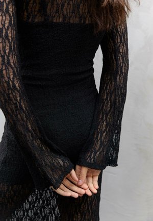 Abito nero in pizzo con maniche lunghe, tessuto strutturato, corpetto aderente e polsini svasati, che mette in mostra un design semi-trasparente ai polsi.