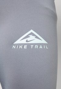 Tissu de sport gris avec un logo triangulaire blanc représentant un éclair et le texte "NIKE TRAIL", texture lisse et design ajusté.