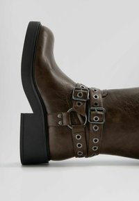 Hnědá kožená kotníková bota s kulatou špičkou, černým klínem a dekorativními pásky s kovovými přezkami a průvleky.