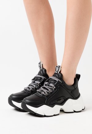 Baskets noires à semelles épaisses blanches, lacets noirs et blancs, portées pieds nus sur un fond blanc.