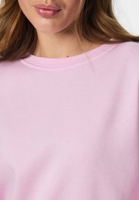 Jeune femme aux longs cheveux bruns portant un t-shirt rose clair à col rond, montrant partiellement ses lèvres et le bas du visage.