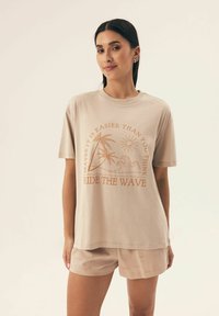 Beżowa bawełniana koszulka z krótkim rękawem, z pomarańczową grafiką palm, słońca i fal. Połączona z pasującymi beżowymi szortami.
