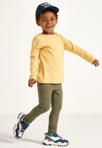 Gul langærmet skjorte, olivengrønne leggings og multicolore sneakers. Barnet bærer en marineblå kasket med teksten "TENNIS KLUB".